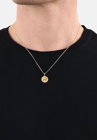 Guldkedja med en rund berlock som har en graverad design. Kedjan är tunn och silverfärgad, bärs över en svart t-shirt med rund hals.