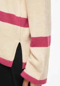 Pull beige avec larges rayures roses sur les manches et le corps, détail fendu sur le côté, porté par-dessus un pantalon noir, gros plan sur le bras et la taille.