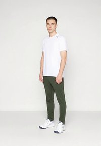 Vīrietis stāv, ģērbies baltā īsā piedurknē T-kreklā, olīvzaļās šaurās biksēs un baltās sporta apavās uz neitrāla gaiši fona.