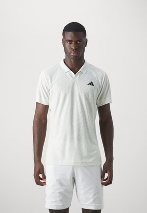 Hellgrünes Poloshirt mit strukturiertem Muster und schwarzem Adidas-Logo auf der linken Brust. Getragen mit weißen Shorts.