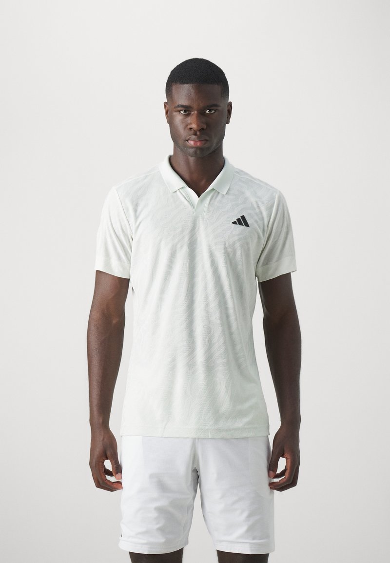 Polo shirt vert clair avec un motif texturé et le logo Adidas noir sur la poitrine gauche. Porté avec un short blanc.