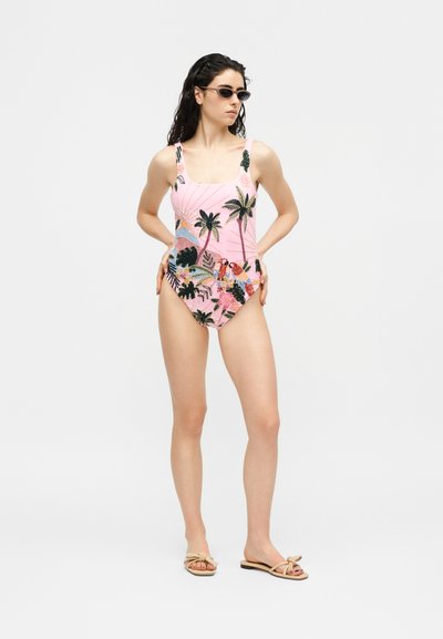 Femme aux cheveux foncés portant un maillot de bain une-pièce rose à imprimé tropical, des lunettes de soleil noires et des sandales beiges, debout les mains sur les hanches.