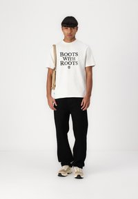 Fehér pamut póló "BOOTS WITH ROOTS" fekete felirattal, fekete nadrággal és világos bézs sportcipővel párosítva. A modell egy világosbarna táskát tart.