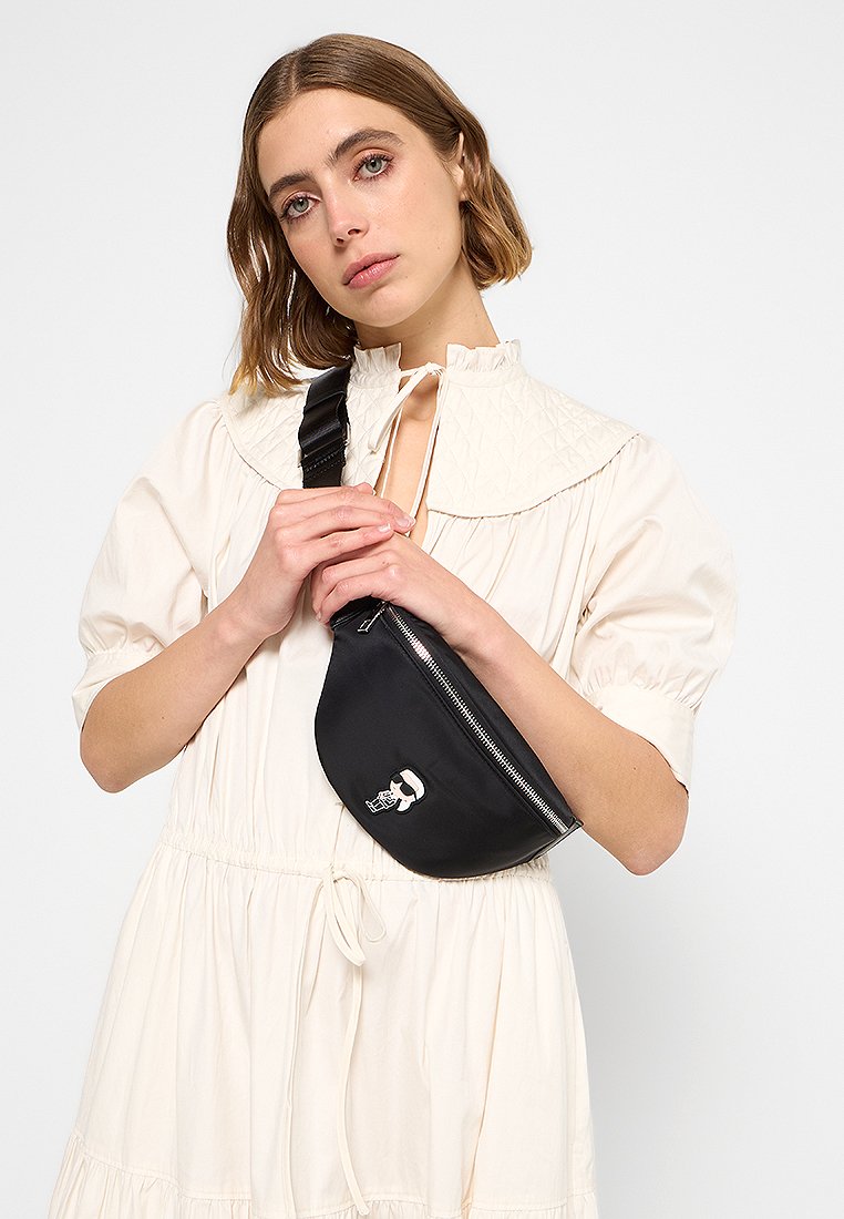 Sac banane en nylon noir avec une forme incurvée, fermeture éclair argentée et un graphique de crâne de dessin animé. Porté en bandoulière avec une sangle réglable.