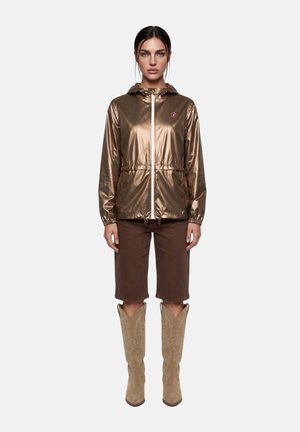 Femme aux cheveux foncés portant une veste à capuche bronze brillante, un short marron à hauteur des genoux, et des bottes de cowboy beige brodées, debout sur un fond clair.