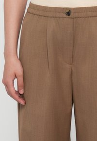 Brune bukser med struktulert stoff, med knappelukking og bretter. Elastisk midje for ekstra komfort.