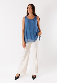 Top senza maniche blu in tessuto trasparente con scollatura rotonda, abbinato a pantaloni bianchi a gamba larga e décolleté neri a punta, su uno sfondo bianco.