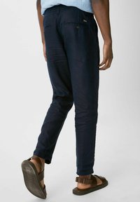 Pantalon marine à coupe slim, fabriqué en tissu léger. Comprend des poches arrière avec fermetures à boutons et des revers retroussés, associé à des sandales marron.