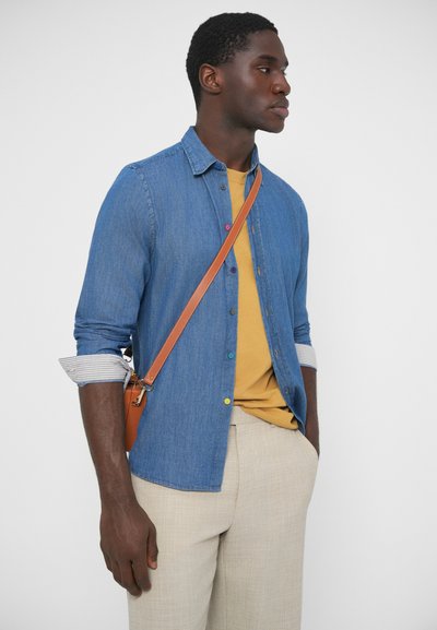 Kék farmer gombos ing, mustár színű pólóval, világos bézs nadrággal és barna oldaltáskával párosítva. Látható csíkos mandzsetta részlet.