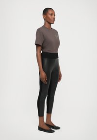 Brown t-shirt with a logo, paired with black high-waisted, fitted leggings. Model wearing black flats, standing side profile against a white backdrop.