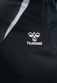 Fekete sportruházat anyagának közelképe, fehér hummel logóval és egy fehér átlós csíkkal a texturált anyagon.
