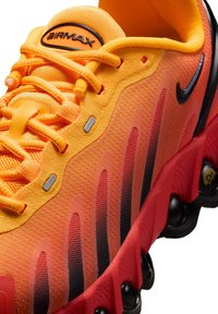A Nike Air Max sportcipők narancs színű hálós felsőrésszel rendelkeznek, fekete és vörös részletekkel, texturált burkolatokkal és a talpban látható légpárnázással.