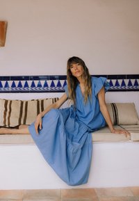 Frau mit langen Haaren, die ein blaues Kleid trägt und auf einer gepolsterten Bank sitzt, mit einer blau-weißen gefliesten Wand im Hintergrund.