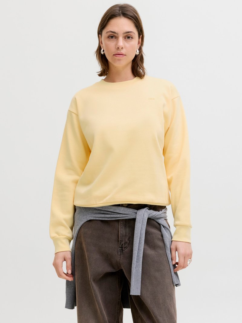 Jeune femme portant un sweat-shirt jaune, un pantalon foncé et un pull gris attaché autour de la taille, posant devant un fond uni.