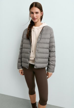 Femme aux cheveux bruns tressés portant une veste matelassée à carreaux sur un sweat à capuche beige et des leggings marron courts, debout contre un mur clair.