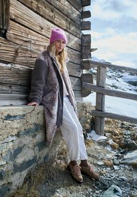 Žena v růžové čepici, vzorovaném koženém kabátu, bílých kalhotách a turistických botách se opírá o dřevěnou chatu s zasněženou horskou krajinou.