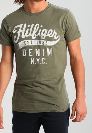 Persoon draagt een olijfgroen T-shirt met witte tekst "Hilfiger Est. 1985 Denim N.Y.C." en een donkere broek, staand tegen een effen achtergrond.