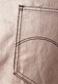 Beiger Denim-Stoff mit einer genähten Taschenbordüre, die mit einer dunklen Fadeneinfassung versehen ist und eine diagonale Struktur auf der Oberfläche aufweist.