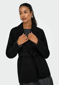 Manteau noir en tissu doux, avec un col cranté, un détail de lien à l'avant et des manches longues, présenté sur un fond clair.