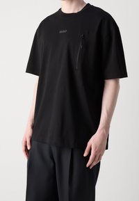 Černé tričko s krátkým rukávem a kapsou na zip na hrudi. Vyrobeno z hladké tkaniny, s jemným logem a strukturovanými švy.