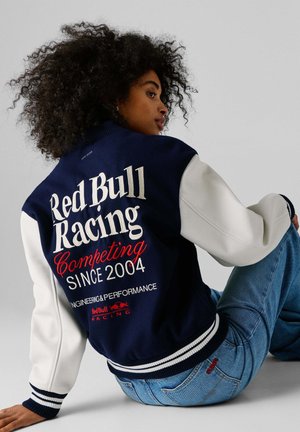 Osoba s kučeravými vlasmi, oblečená v námornícko-modro-bielej bundě typu varsity s nápisom "Red Bull Racing" a modrých džínsoch, sediaca na zemi.