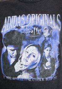 Melnā T-kreklā ir nolietota grafika ar "Buffy pret Vampīriem" varoņiem zilās krāsās, ar tekstu: "Adidas Originals" un "Ja apokalipse nāk...".