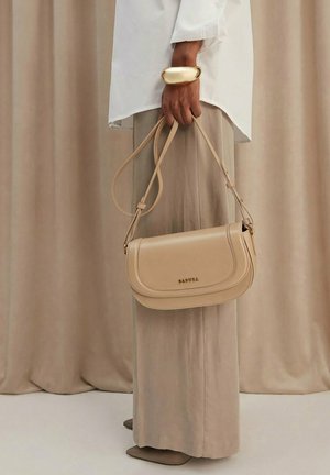 Mano sosteniendo un bolso de hombro Badura de cuero beige, con blusa crema, pantalones de pierna ancha, brazalete dorado, de pie frente a un fondo de cortina color coñac.