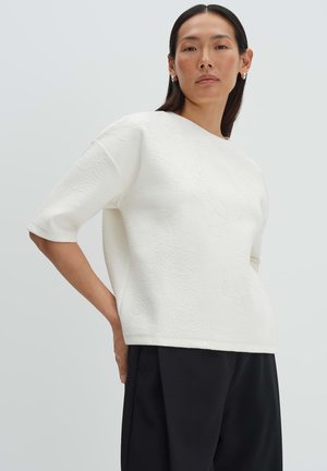 Frau trägt ein weißes, strukturiertes Kurzarm-Top mit Blumenmuster und schwarze, weit geschnittene Hosen, steht mit einer Hand in der Hüfte.