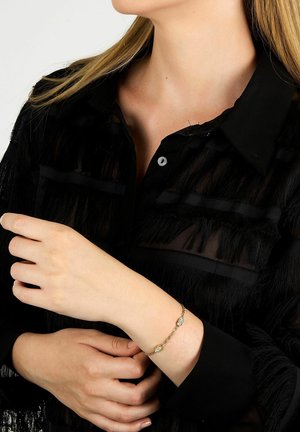 Guldtonet armbånd med flere ovale accenter, båret på et håndled. Sort, frynset stofskjorte delvist synlig i baggrunden.