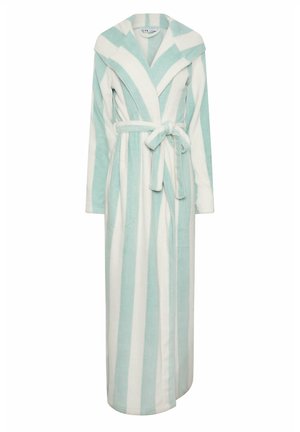 Femme portant un long peignoir à capuche avec des rayures verticales blanches et bleu clair, mains dans les poches, debout devant un fond uni.