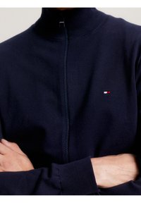 Suéter azul marino con cierre, hecho de una tela suave. Presenta un cuello alto, puños acanalados y un pequeño logotipo en rojo, blanco y azul en el pecho.