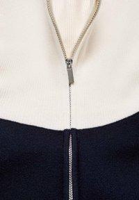 Pull en tricot avec une demi-fermeture éclair. Comprend une partie supérieure crème et une partie inférieure bleu marine. Tissu texturé avec détails métalliques sur la fermeture éclair.