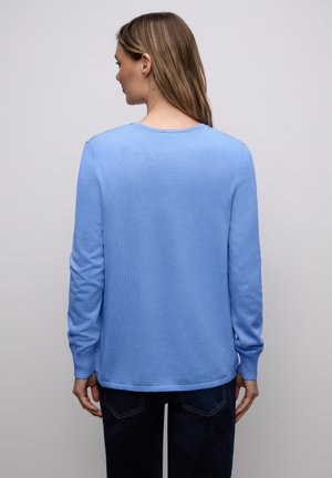 Femme vue de dos portant un pull bleu clair à manches longues et un jean foncé sur un fond uni gris clair.