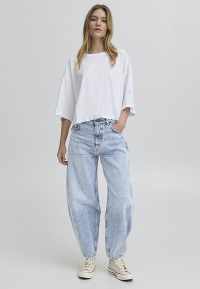 Bijela skraćena majica s nisko postavljenim ramenima, uparena s labavim, svijetloplavim trapericama. Traperice imaju izblijedjeli izgled i sužene rubove.