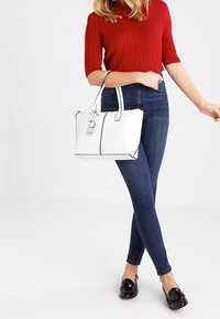 Witte handtas met zwarte accenten, gestructureerd ontwerp en twee bovenhandvatten, gecombineerd met strakke donkerblauwe jeans en een rode geribde trui.