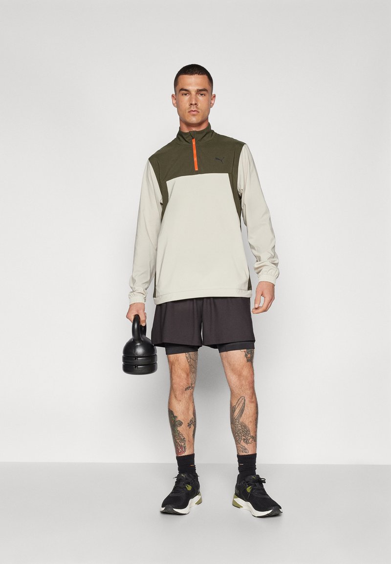 Man in een gekleurde langmouwen sporttop en zwarte shorts die een zwarte kettlebell vasthoudt, staand tegen een effen lichte achtergrond.