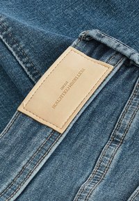 Tissu en denim d'une teinte bleue moyenne. Une étiquette en cuir marron sur la ceinture indique "Next Denim Collection Established 1992." Détails de couture visibles.