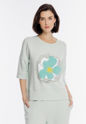 Kobieta w jasnopzielonej bluzie z dużym wzorem niebiesko-żółtego kwiatu, stojąca z rękami w kieszeniach na białym tle.