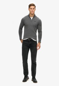Golfino grigio con zip e finitura texture, indossato sopra una camicia bianca, abbinato a jeans neri e scarpe nere stringate. Look informale.
