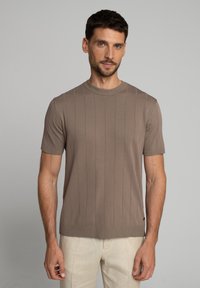 Mężczyzna z krótkimi ciemnymi włosami, ubrany w taupe'owy, prążkowany sweter z krótkim rękawem i jasnobeżowe spodnie, na jednolitym szarym tle.