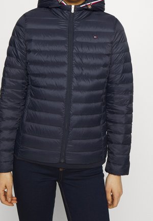 Personne portant une doudoune matelassée bleu marine zippée avec capuche et petit logo sur la poitrine, associée à un pantalon foncé ajusté.