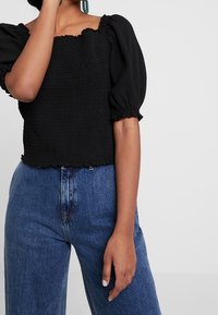 Černý top s čtveratým výstřihem, texturovaným smockovaným vzorem, balónovými rukávy a elastickým lemem, spárovaný s modrými džíny s vysokým pasem.