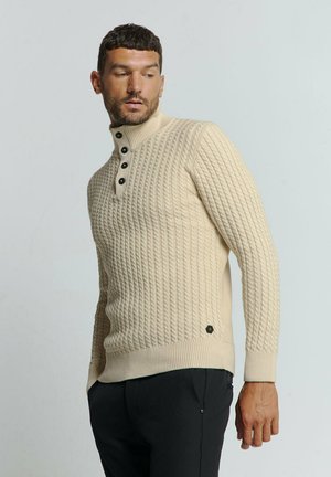 Beige gebreide trui met een hoge kraag, vier zwarte knopen, geribbelde manchetten en een klein logoaccent aan de zoom.