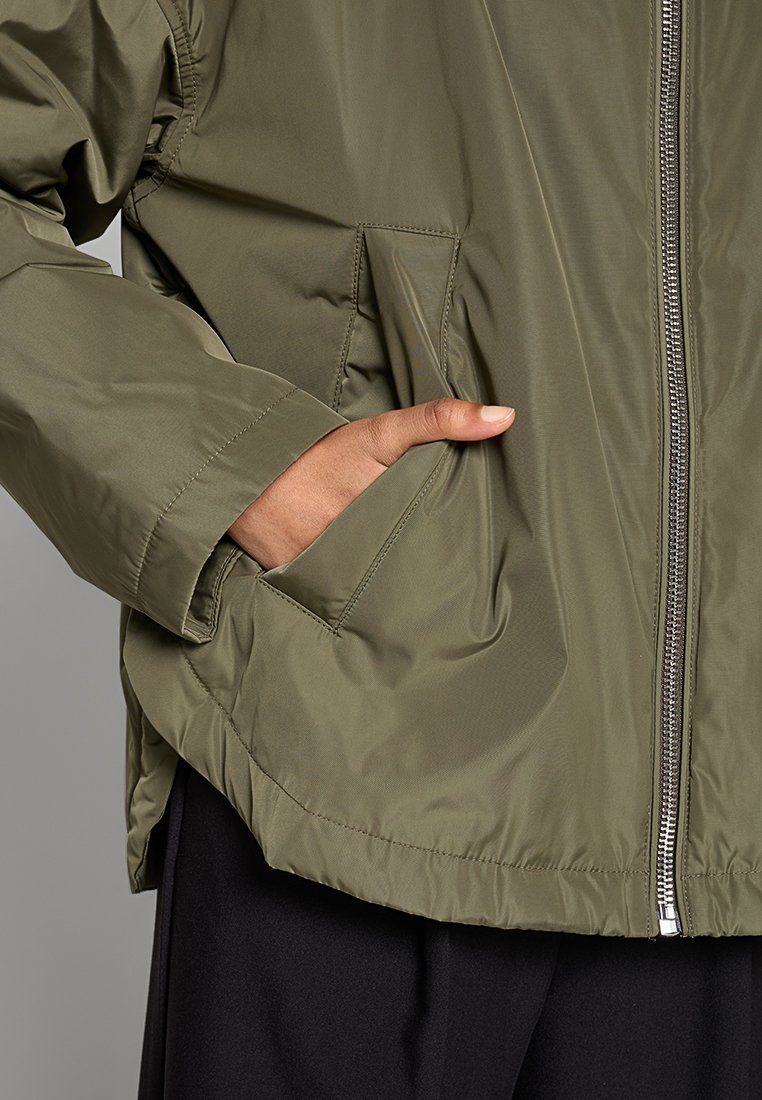 Olivgrüne Jacke mit Reißverschluss, seitlichen Taschen und einer glatten, matten Textur. Maßgeschneiderter Schnitt mit einer lässigen Passform.