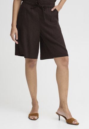 Nő sötétbarna, térdig érő rövidnadrágban és bézs színű, nyitott orrú magassarkú szandálban, zsebbe tett kézzel, fehér háttér előtt áll.