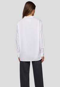 White long-sleeve shirt with a collar, featuring a smooth texture and a relaxed fit. Shown from the back, tucked into dark trousers.
