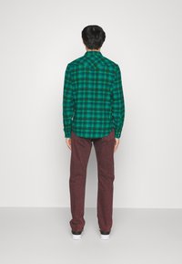 Hombre de pie de espaldas, vistiendo una camisa de cuadros verde y azul, jeans burdeos y zapatillas blancas, contra un fondo blanco liso.