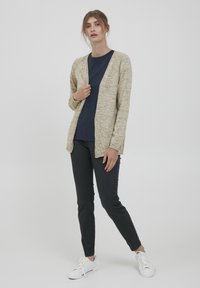 Cardigan en tricot beige avec un devant ouvert, des manches longues et des poignets côtelés, superposé sur un haut bleu marine, associé à un pantalon noir et des baskets blanches.