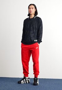 Camisa preta de mangas longas com padrão tonal, calças de moletom vermelhas e ténis pretos com detalhes brancos. Figura independente sobre uma superfície plana.