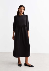 Black, long-sleeve dress with a textured fabric, a round neckline, and gathered waist. Paired with black flat shoes. Simple, ankle-length design.